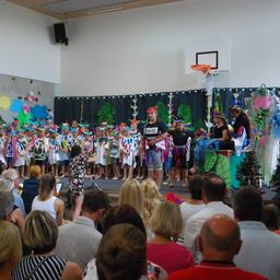 Eine Gruppe von Kindern auf der Bühne, in farbenfrohen Kostümen, tritt vor einem Publikum auf. Hinter ihnen sind schwarze Vorhänge mit Dekorationen und einem Basketballkorb.