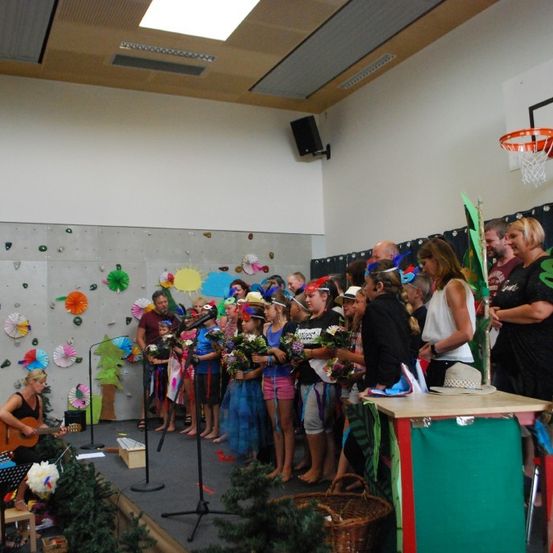 Eine Gruppe von Kindern und Erwachsenen versammelt sich auf einer Bühne mit einer Frau, die Gitarre spielt, während andere Blumen halten und Hüte tragen. Der Raum hat eine Kletterwand und einen Basketballkorb.