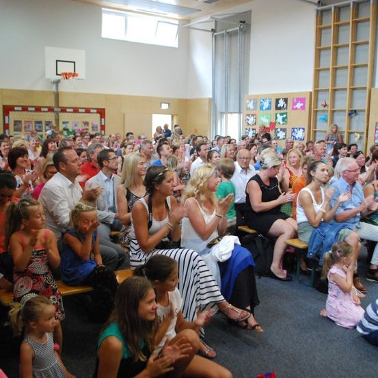 Eine große Gruppe von Menschen versammelt sich in einem Raum, einige stehen, andere sitzen auf Bänken. Sie scheinen zu klatschen und zu jubeln. Der Raum hat weiße Wände und einen Basketballkorb.