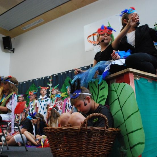 Kinder in Kostümen führen ein Theaterstück in einem Klassenzimmer mit Bühne und Mikrofon-Setup auf. Ein Kind sitzt in einem Korb, während andere zuschauen.