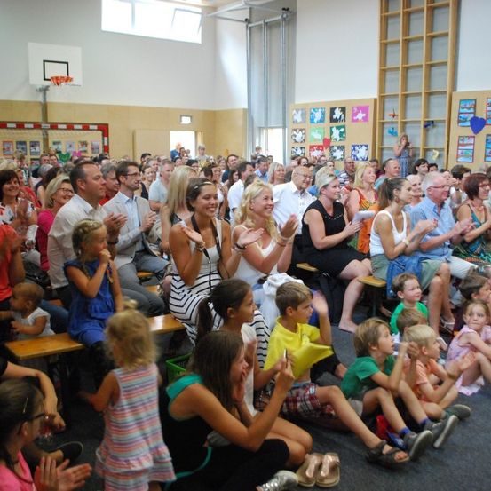 Eine große Gruppe von Menschen sitzt und steht in einer Turnhalle und klatscht. Einige sitzen auf Bänken, andere stehen in der Nähe von Wänden mit Kunstwerken. Kinder sind unter den Anwesenden.