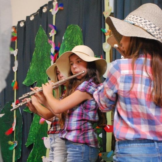 Drei Mädchen in Cowboyhüten spielen Flöten vor einem Hintergrund mit grünen Bäumen und bunten Dekorationen.