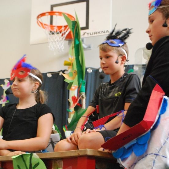 Drei Kinder mit bunten Masken und Kopfhörern sitzen auf einer Bank. Sie nehmen wahrscheinlich an einem Schultheaterstück oder einer Aufführung teil.