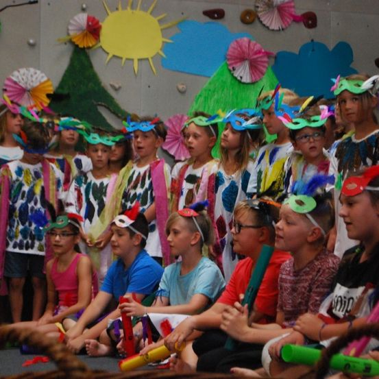 Eine Gruppe von Kindern in Kostümen und Masken sitzt in einer Reihe auf der Bühne und hält Musikinstrumente. Bunte Dekorationen schmücken den Hintergrund.