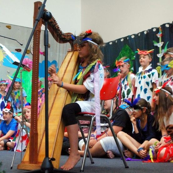 Ein Mädchen in einem bunten Kostüm spielt eine Harfe auf der Bühne, während eine Gruppe von Kindern vor ihr sitzt, die Masken und Kostüme tragen.