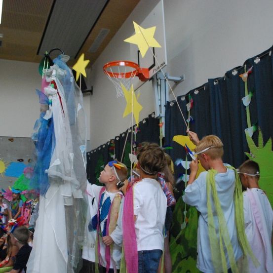 Kinder in Kostümen mit Engelsflügeln nehmen an einer festlichen Veranstaltung in einem mit bunten Dekorationen geschmückten Raum teil.