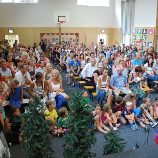 Eine große Menschenmenge, darunter auch Kinder, sitzt auf Bänken in einer Turnhalle. Im Hintergrund stehen mehrere Erwachsene, und im Vordergrund befinden sich zwei künstliche Bäume.