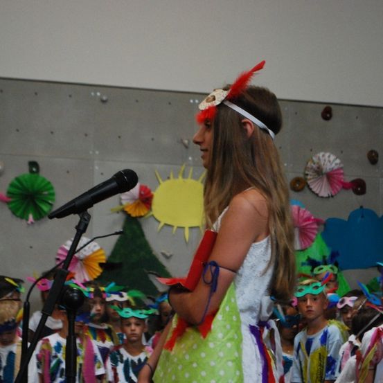 Ein junges Mädchen mit einem Kopfschmuck steht vor einem Mikrofon, umgeben von Kindern in Kostümen, und führt eine Aufführung in einem dekorierten Raum durch.