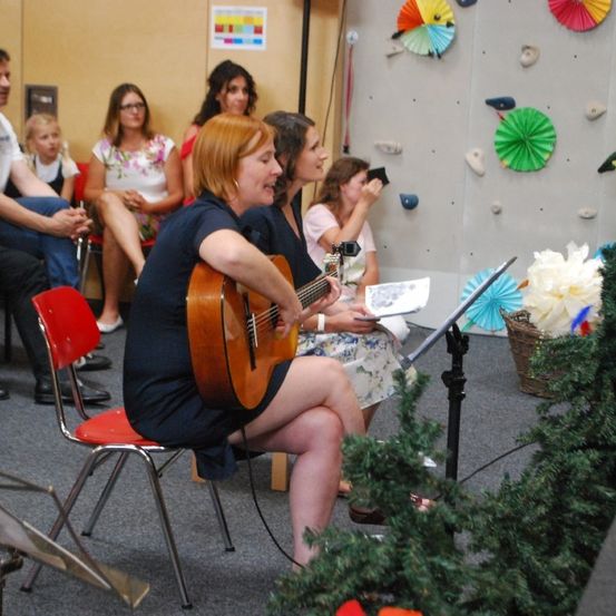 Eine Frau spielt Gitarre, während sie auf einem Stuhl vor einer Wand mit Klettergriffen sitzt. Andere schauen zu.