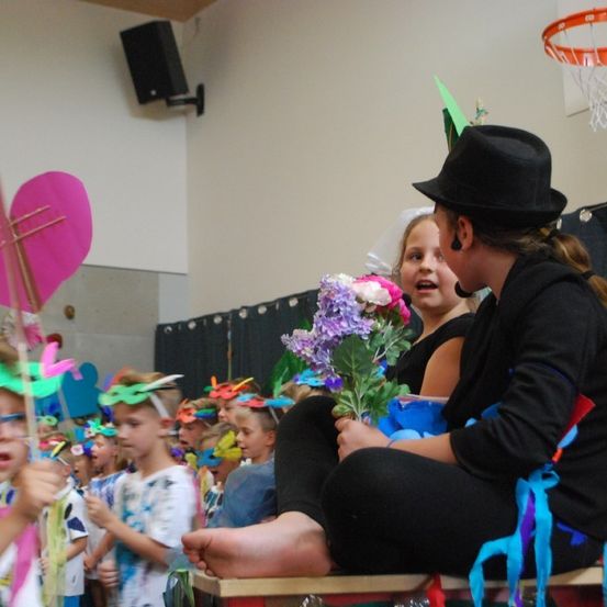 Eine Gruppe von Kindern in einem Klassenzimmer ist in Partyhüten gekleidet und hält Blumen, während eine Frau in einem Hut auf der Bühne sitzt.