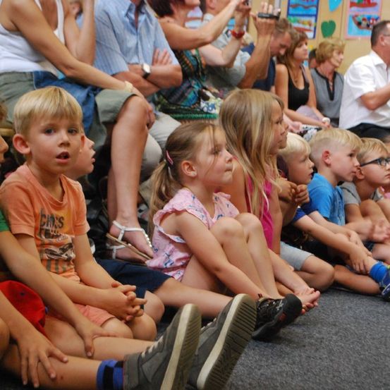 Eine Gruppe von Kindern und Erwachsenen sitzt auf dem Boden. Die Kinder schauen nach oben, während die Erwachsenen klatschen. Einige der Kinder tragen Brillen.