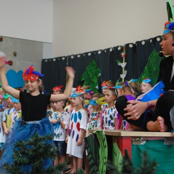 Eine Gruppe von Kindern in bunten Kostümen und Masken führt eine Aufführung im Klassenzimmer durch. Ein Kind leitet die Aufführung auf der Bühne.
