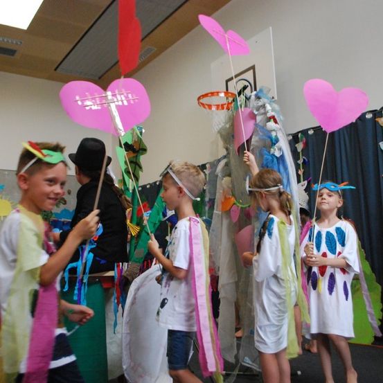 Eine Gruppe von Kindern in bunten Kostümen, die rosa herzförmige Objekte halten, steht in einem Raum mit grünem Hintergrund und Dekorationen.