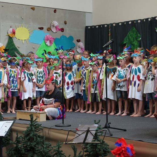 Eine Gruppe von Kindern in farbenfrohen Kostümen und Masken tritt auf einer Bühne mit einem Hintergrund aus bunten Dekorationen auf.