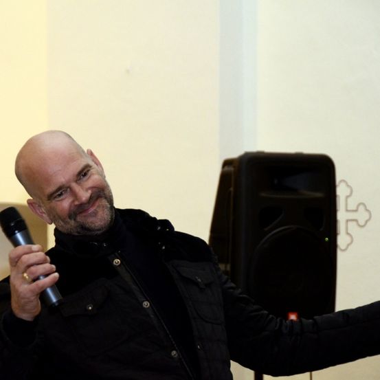 Ein bärtiger Mann lächelt und hält ein Mikrofon. Er trägt einen schwarzen Mantel und einen Ring. Hinter ihm steht ein Lautsprecher.