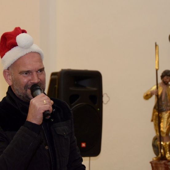 Ein Mann mit einem Weihnachtshut spricht in ein Mikrofon, mit einem Lautsprecher hinter ihm und einer goldenen Statue zu seiner Rechten.