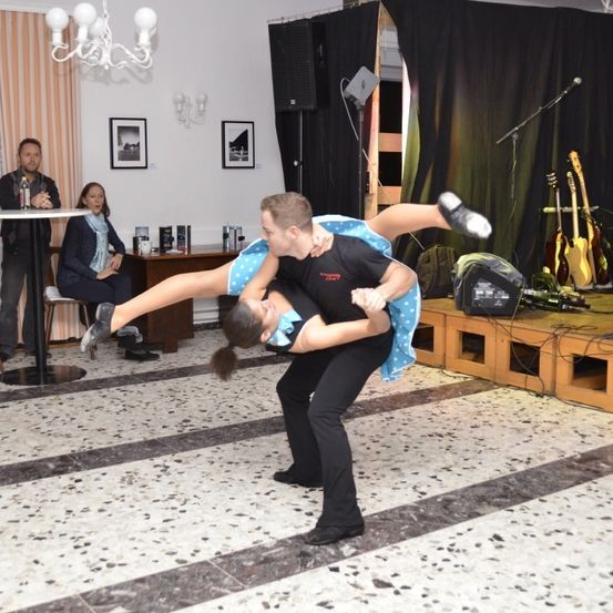 Ein Paar tanzt in einem Raum mit gefliestem Boden, während andere sitzen und zusehen. Eine Kronleuchter hängt über ihnen. In der Nähe befinden sich Musikinstrumente.