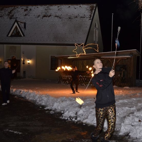 Drei Personen führen einen Feuertanz vor einem Gebäude mit einem sternförmigen Licht auf dem Dach auf. Sie stehen auf einem verschneiten Boden.