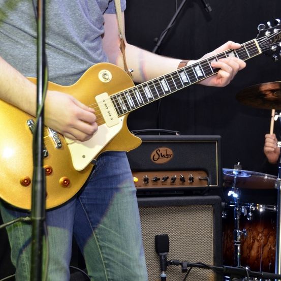 Eine Person steht und hält eine goldene E-Gitarre, trägt ein graues Shirt und Jeans und steht vor einem schwarzen Verstärker und Schlagzeug mit Becken.
