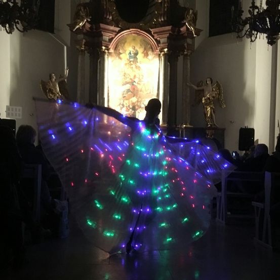 Eine Person tanzt in einer Kirche mit einem Kleid, das von farbigen Lichtern beleuchtet wird, während Menschen auf den Kirchenbänken zusehen.