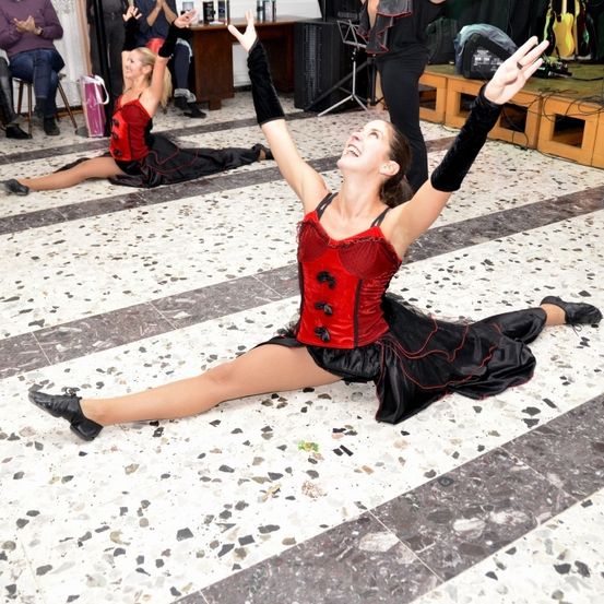 Zwei Ballerinen führen auf einer Bühne auf, eine sitzt, die andere kniet, beide tragen rote und schwarze Kostüme. Sie spielen vor einem Publikum mit Musikinstrumenten auf einer hölzernen Bühne.