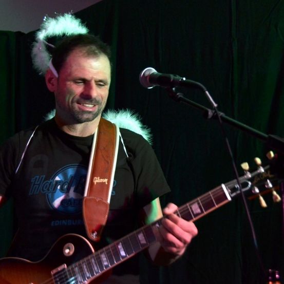 Ein Mann spielt eine E-Gitarre auf der Bühne. Er trägt ein schwarzes T-Shirt mit einem Hard Rock Cafe Logo. Ein Mikrofon steht vor ihm.