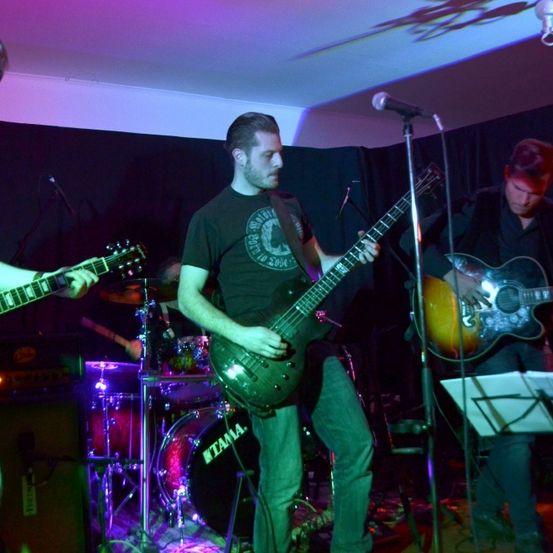 Eine Band spielt auf der Bühne mit grünem und lila Licht. Der Leadgitarrist spielt eine E-Gitarre, während andere Bass und Schlagzeug spielen. Mikrofone und Lautsprecher sind auf der Bühne.