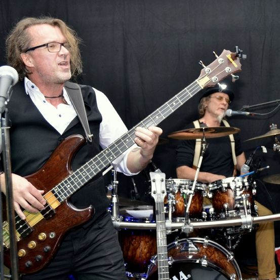 Ein Mann in einem schwarzen Weste spielt eine Bassgitarre und singt in ein Mikrofon. Ein anderer Mann spielt Schlagzeug hinter ihm. Sie sind auf der Bühne mit einem schwarzen Hintergrund.