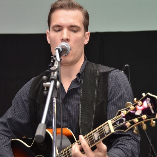 Ein Mann tritt auf der Bühne auf, singt in ein Mikrofon und spielt eine akustische Gitarre. Er trägt eine schwarze Weste und ein schwarzes Hemd.