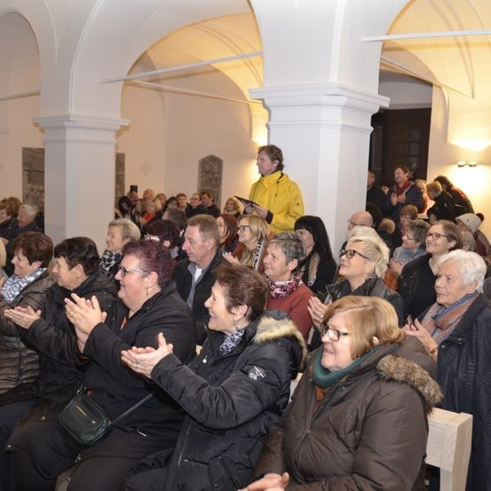 Eine große Gruppe von Menschen, hauptsächlich ältere Menschen, sitzt in einer Kirche, klatscht und lächelt. Ein Mann in einer gelben Jacke steht im Hintergrund.