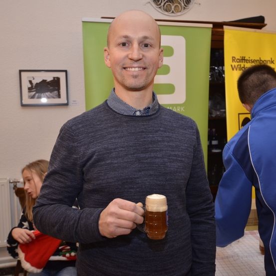 Ein Mann in einem grauen Pullover hält ein Bierglas und lächelt in die Kamera. Hinter ihm sind ein Banner und ein Bilderrahmen zu sehen. Eine Frau und ein Kind sind ebenfalls in der Szene, mit einem Heizkörper im Hintergrund.