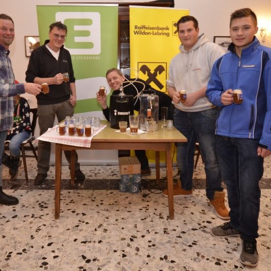 Eine Gruppe von Menschen steht um einen Tisch mit Biergläsern. Hinter ihnen sind zwei Banner sichtbar, eines mit einem Logo und Text, und ein anderes mit einem 'X'. Einige Personen sitzen auf Stühlen.