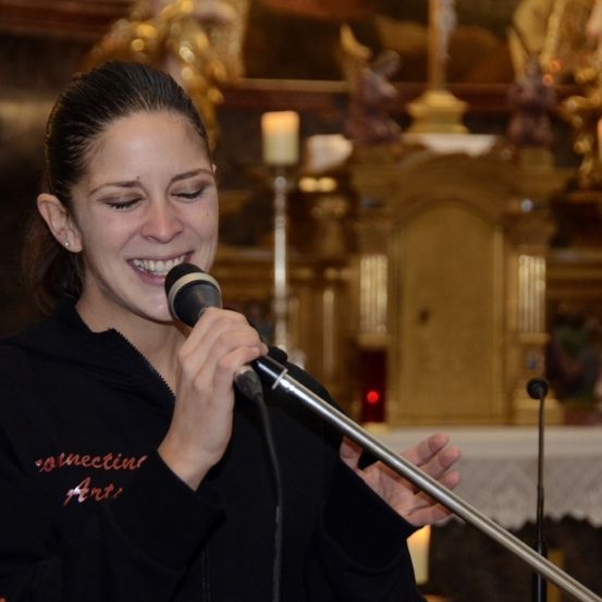 Eine Frau singt in ein Mikrofon in einer Kirche, mit Kerzen und einem verzierten Altar hinter ihr. Sie trägt eine schwarze Sweatshirtjacke mit rotem Schriftzug.