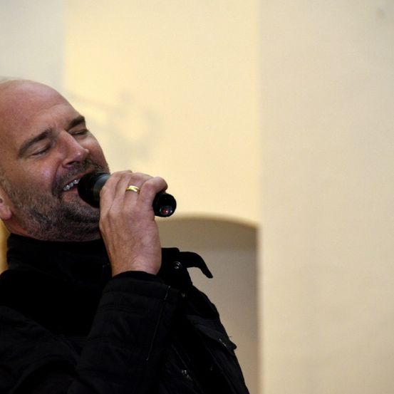 Ein kahlköpfiger Mann mit Bart singt in ein Mikrofon, die Augen geschlossen, in einem Raum mit gelber Wand.