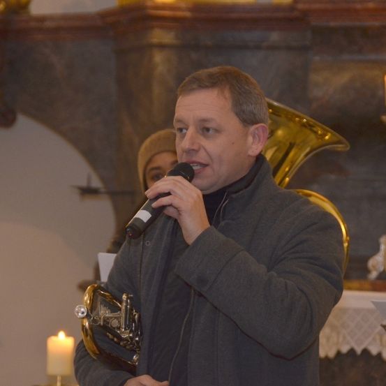 Ein Mann spricht in ein Mikrofon und hält ein Blechblasinstrument. Eine Frau steht hinter ihm, und zwei brennende Kerzen sind vorhanden. Eine Tuba ist auf der rechten Seite, und eine Steinmauer ist im Hintergrund.