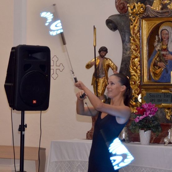 Eine Frau führt mit beleuchteten Stäben in einer Kirche auf, mit einer Statue, einem Gemälde und Blumen im Hintergrund.