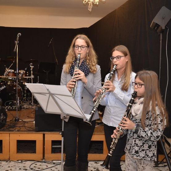 Drei junge Mädchen, alle mit Brille, spielen Klarinetten auf einer Bühne. Dahinter ist ein Drum-Set und andere Instrumente aufgebaut.