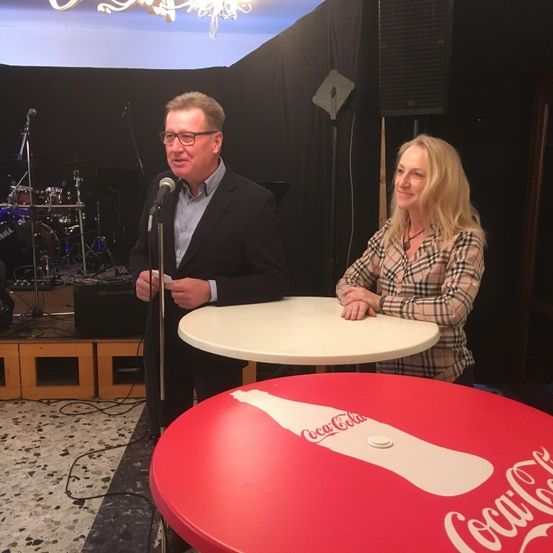 Ein Mann spricht in ein Mikrofon, während eine Frau zusieht, beide sitzen an einem Tisch. Dahinter befindet sich eine Bühne mit Trommeln und Lautsprechern. Der Tisch zeigt ein Coca-Cola-Logo.