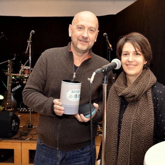 Ein Mann und eine Frau stehen lächelnd vor einem Mikrofon. Der Mann hält eine Tasse und einen kleinen Lautsprecher. Im Hintergrund sind Musikinstrumente zu sehen.