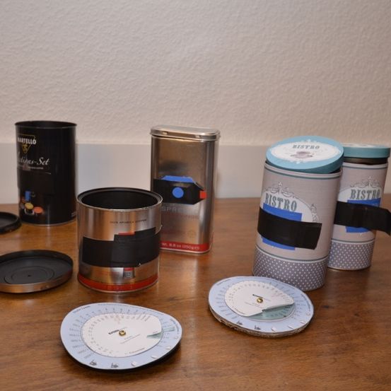 Auf einem Holztisch sind zylindrische Behälter ausgestellt, darunter eine schwarze zylindrische Dose, eine silberne Dose mit der Aufschrift Spresso und zwei zylindrische Dosen mit der Aufschrift Bistro. Zusätzliche Gegenstände sind runde Scheiben und ein kreisförmiges Messwerkzeug.