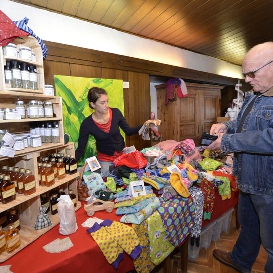 Ein älterer Mann betrachtet verschiedene Gegenstände auf einem Tisch, während eine Frau ihm hilft, umgeben von Gläsern, Textilien und einem Holzschrank.