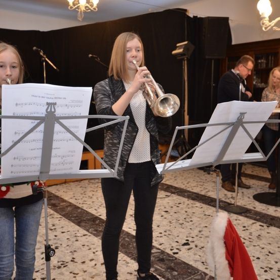 Zwei junge Mädchen spielen Trompeten bei einem Konzert. Sie haben Notenständer vor sich mit Musikblättern. Dahinter stehen ein Mann und eine Frau. Es gibt Mikrofone und Lautsprecher auf Ständern.