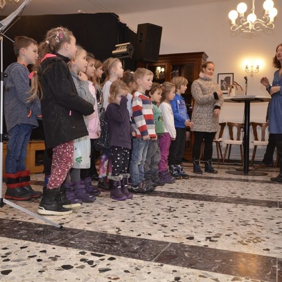 Eine Gruppe von Kindern in Winterkleidung steht in einem Raum mit einer Frau, wahrscheinlich einer Lehrerin, die an einem Podium spricht. Eine Kronleuchter und ein Bilderrahmen schmücken die Wände.