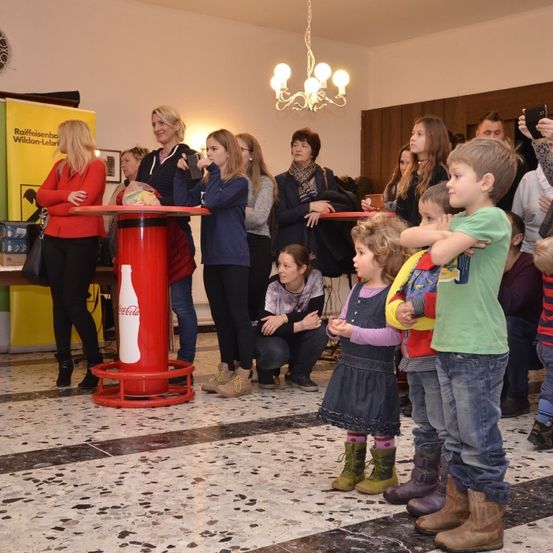 Eine Gruppe von Menschen ist um eine rote Coca-Cola-Flasche in einem Raum mit weißen Wänden versammelt. Einige halten Tassen und machen Fotos, während andere beobachten. Der Raum ist gut beleuchtet mit einer Kronleuchter.