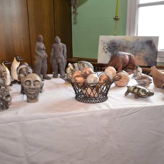 Ein Tisch mit verschiedenen Skulpturen, darunter menschliche Gesichter, Tiere und Eier in einem Korb, vor dem Hintergrund eines Fensters und einer Kerze.