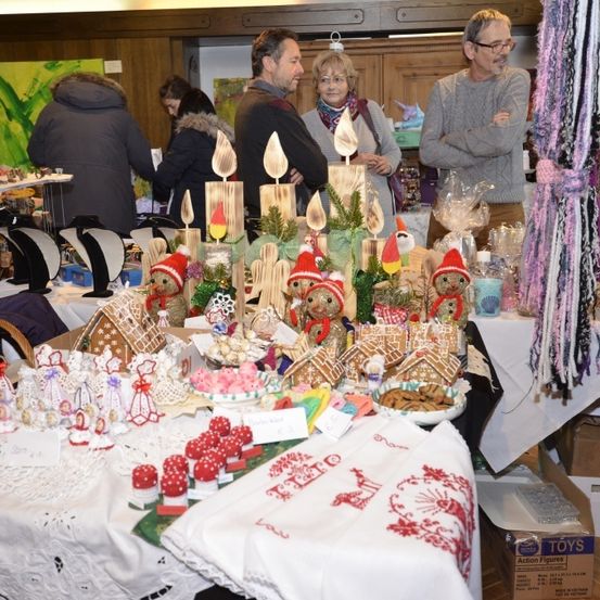 Eine Gruppe von Menschen versammelt sich um einen Tisch, der mit verschiedenen Lebkuchenhäusern, Keksen und Dekorationen gefüllt ist. Sie stehen hinter dem Tisch und einige betrachten die Gegenstände. Der Tisch ist mit einem weißen Tuch bedeckt und es gibt zusätzliche Gegenstände an der Seite.