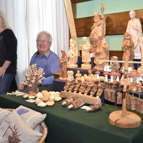 Ein Tisch zeigt verschiedene Holzarbeiten, mit einem Mann und einer Frau dahinter, die lächeln. Verschiedene geschnitzte Figuren und Gegenstände sind auf dem Tisch, zusammen mit einem weißen Vorhang im Hintergrund.