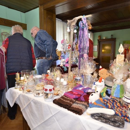 Zwei ältere Menschen betrachten einen Tisch voller verschiedener Gegenstände. Einer trägt eine Schürze und der andere eine Jeansjacke. Hinter ihnen befindet sich eine grüne Wand mit einem Fenster.