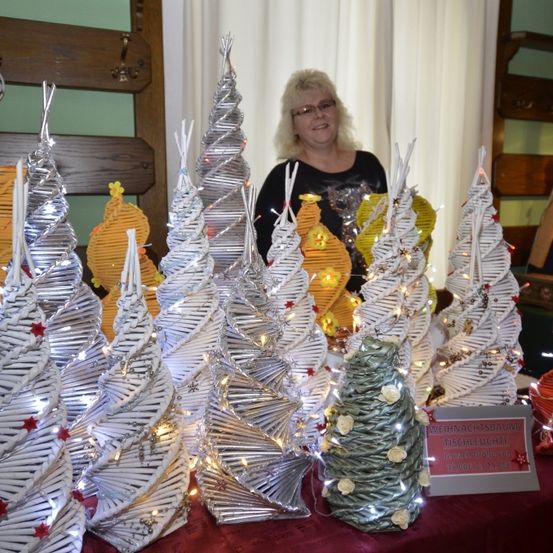 Eine Frau steht hinter einer Ausstellung von spiralförmigen Weihnachtsbäumen, einige mit Lichtern und Ornamenten geschmückt. Die Bäume sind in verschiedenen Farben und Größen, mit einem Schild in Deutsch in der Nähe.