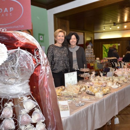 Zwei Frauen stehen hinter einem Tisch, auf dem Produkte von Soap Stars ausgestellt sind. Der Tisch ist mit verschiedenen Seifen und Geschenken beladen. Eine Frau trägt ein schwarz-weißes Oberteil, die andere ein schwarzes Sweatshirt.